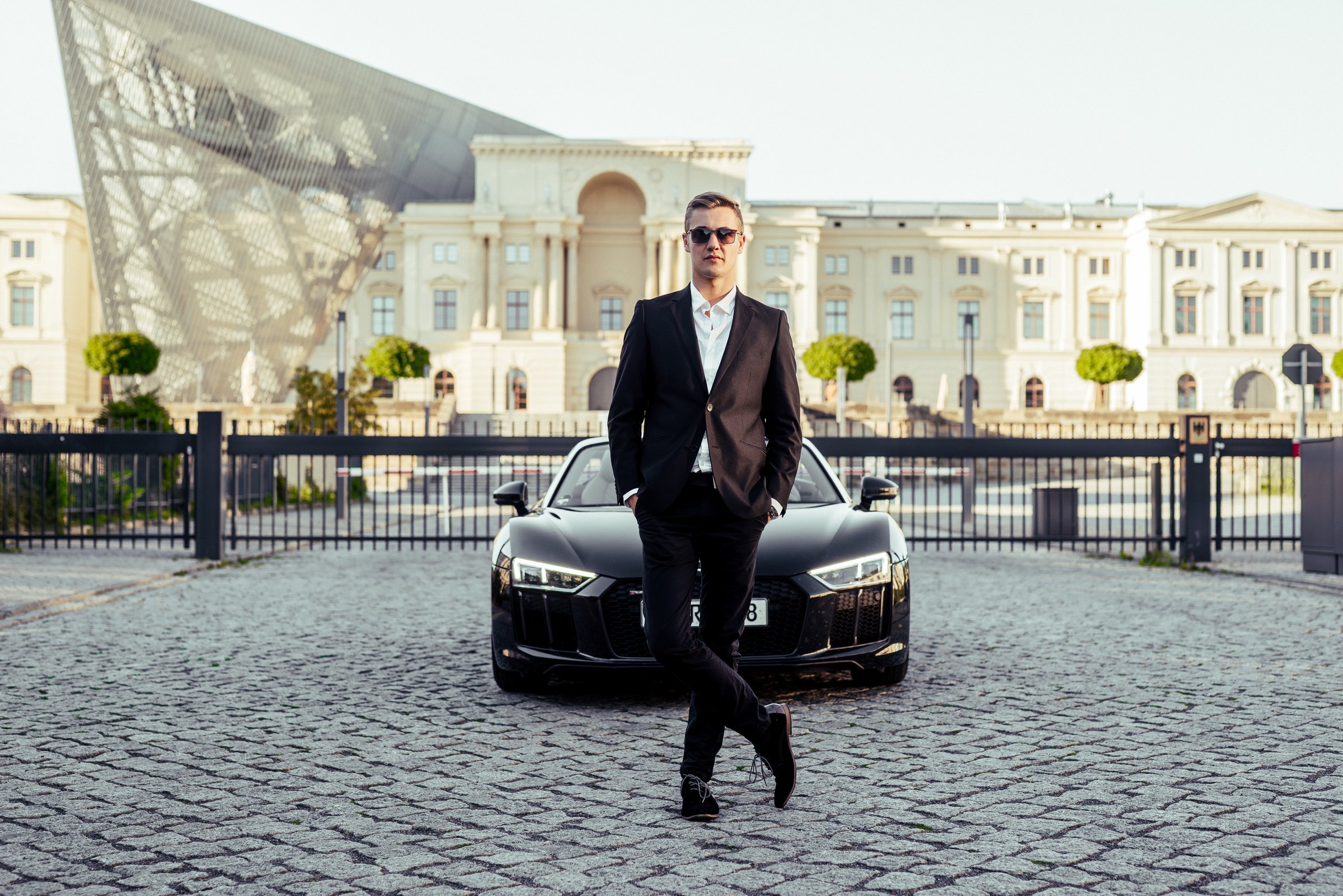 Max Haase in a black suit in front of an Audi R8 Spyder on cobblestones