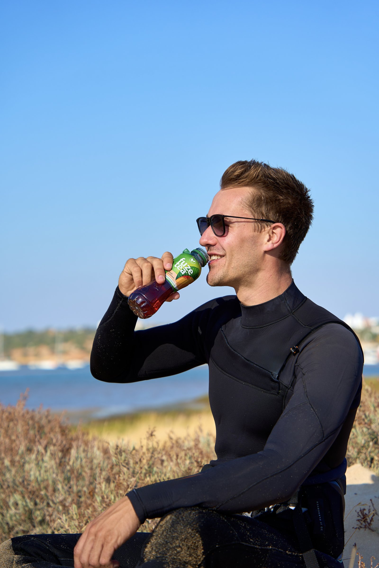 Max Haase, wearing a wetsuit, drinks from a Fuze Tea bottle by the water.