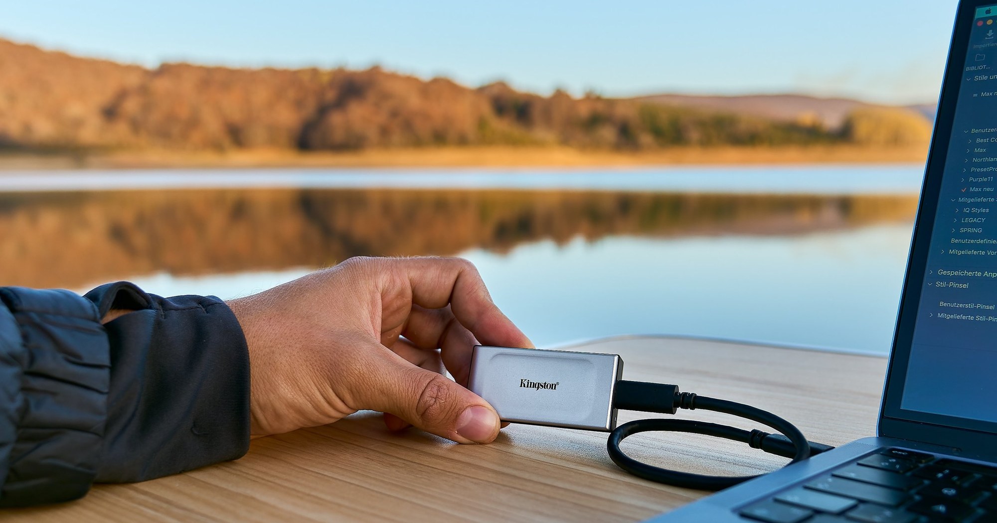 A hand holds a Kingston SSD next to an open laptop in front of a lake.